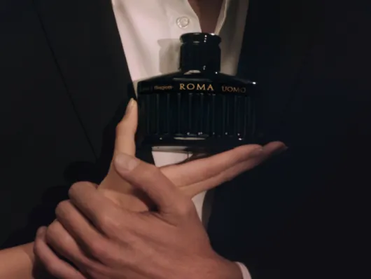 Mano maschile ed elegante che sorregge il flacone del profumo Roma Uomo di Laura Biagiotti, con uno sfondo scuro e una giacca formale che suggeriscono un'atmosfera sofisticata.